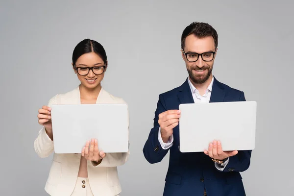 Міжрасова пара бізнес-колег в окулярах тримає ноутбуки ізольовані на сірому — стокове фото
