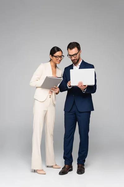 Full length view of interracial business colleagues with laptop and folder on grey — Stock Photo