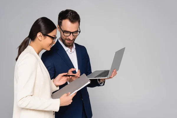 Bearded businessman pointing with finger at folder in hands of asian colleague while holding laptop isolated on grey — Stock Photo