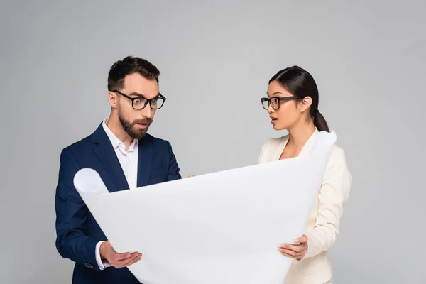 Asian businesswoman looking at shocked business colleague while holding white paper isolated on grey — Stock Photo