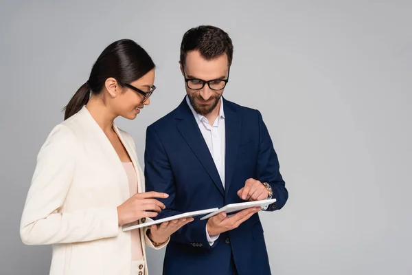Interracial couple of businesspeople in formal wear and eyeglasses using digital tablets isolated on grey — Stock Photo