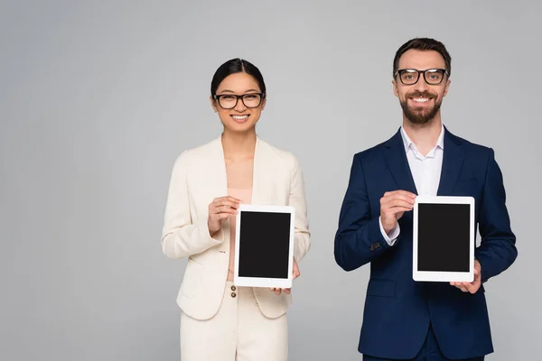 Міжрасові пари бізнес-партнерів в окулярах, що показують цифрові планшети з порожнім екраном ізольовані на сірому — стокове фото
