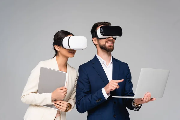 Mujer de negocios sosteniendo la carpeta mientras usa auriculares vr junto con su colega apuntando con el dedo a la computadora portátil aislada en gris - foto de stock