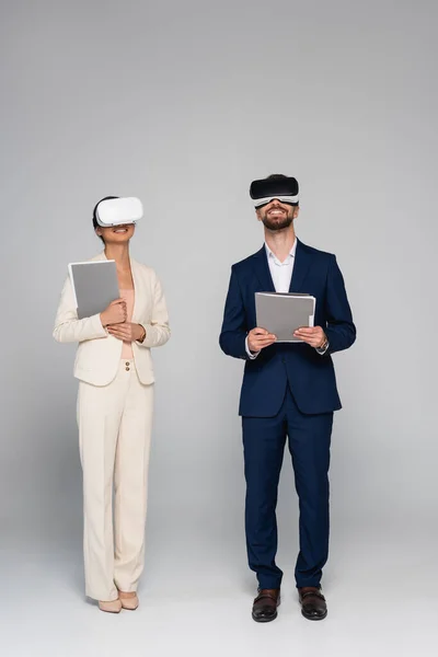 Full length view of businesspeople in suits and vr headsets holding documents while standing on grey — Stock Photo
