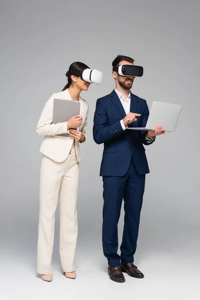 Full length view of businessman in vr headset pointing with finger at laptop while using vr headsets together with colleague on grey — Stock Photo