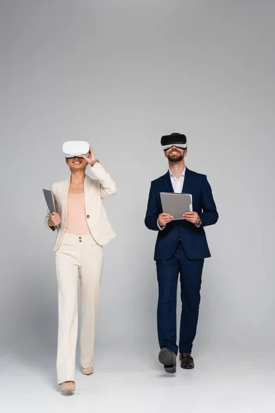 Full length view of business partners in vr headsets holding folders while walking on grey — Stock Photo