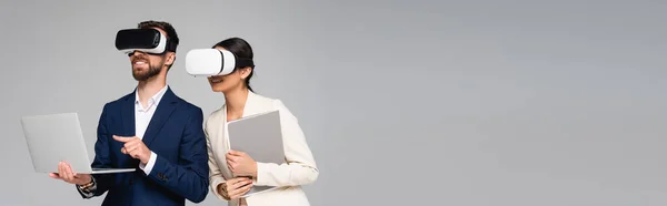 Imagen horizontal del hombre de negocios apuntando con el dedo a la computadora portátil mientras usa auriculares vr junto con su colega aislado en gris - foto de stock