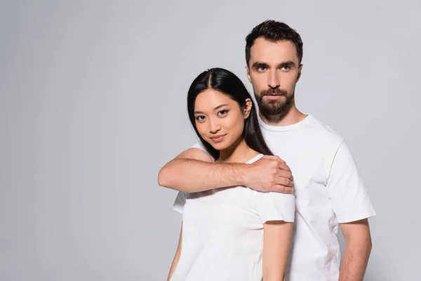 Jovem homem no branco t-shirt abraçando asiático mulher enquanto posando isolado no cinza — Fotografia de Stock