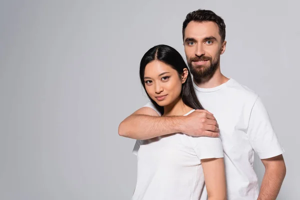 Bearded man in white t-shirt embracing young asian woman while posing isolated on grey — Stock Photo