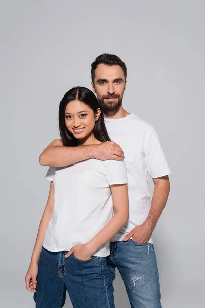 Homem barbudo abraçando morena mulher asiática enquanto posando com as mãos em bolsos isolados no cinza — Stock Photo