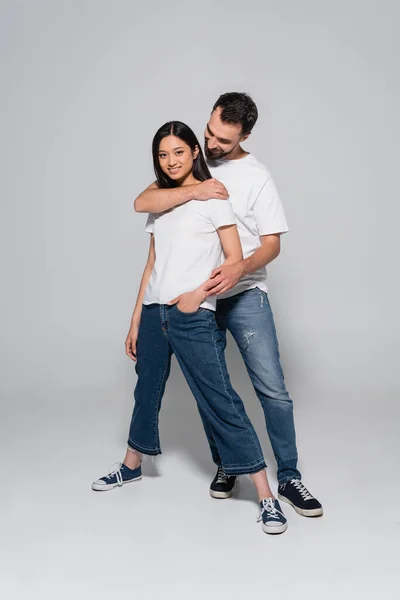 Vista completa del hombre en camiseta blanca y jeans abrazando novia asiática de pie con la mano en el bolsillo en gris - foto de stock