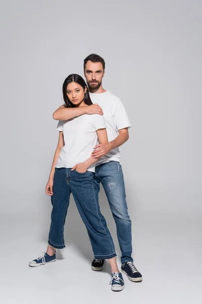 Full length view of confident man in white t-shirt and jeans hugging asian girlfriend while posing on grey — Fotografia de Stock