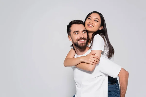 Joven, barbudo hombre en blanco camiseta piggybacking morena asiático novia aislado en blanco - foto de stock