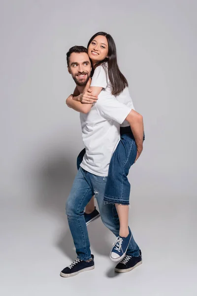 Bärtiger Mann in weißem T-Shirt und Jeans huckepack asiatische Freundin auf grau — Stockfoto