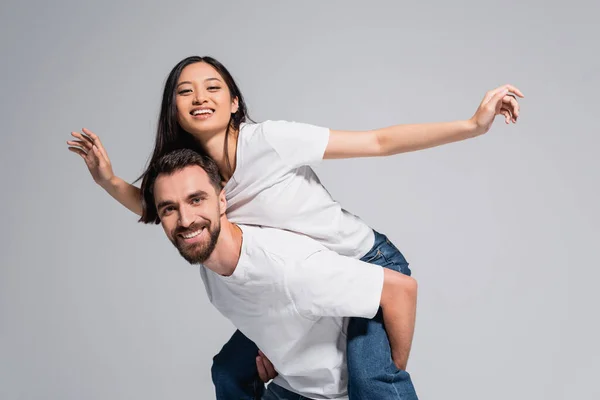 Молодой человек в белой футболке, подпирающий взволнованную азиатскую подружку, имитирующую полет изолированного на сером — стоковое фото