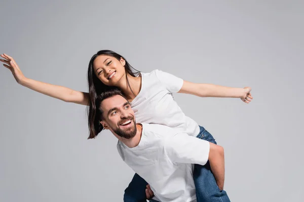 Excited man in white t-shirt piggybacking asian girlfriend imitating flying isolated on grey — Stock Photo