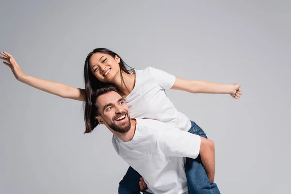 Animado homem no branco t-shirt piggybacking ásia namorada imitando voando isolado no cinza — Fotografia de Stock