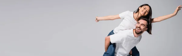 Panoramic orientation of excited man piggybacking asian girlfriend imitating flying isolated on grey — Stock Photo