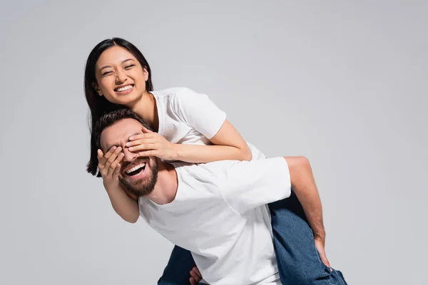 Laughing man piggybacking brunette asian girlfriend covering his eyes with hands isolated on grey — Stock Photo