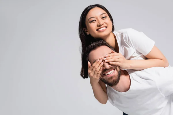 Excited man piggybacking asian girlfriend covering his eyes with hands isolated on grey — Stock Photo