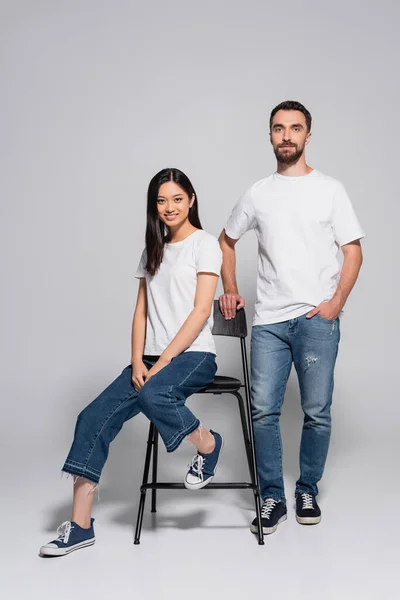 Brünette asiatische Frau in weißem T-Shirt und Jeans sitzt auf Stuhl neben stilvollem Freund stehend mit der Hand in der Tasche auf grau — Stockfoto