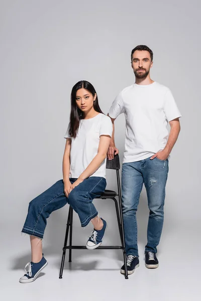 Стильна азіатська жінка в білій футболці і джинсах сидить на стільці біля хлопця, що стоїть з рукою в кишені на сірому — стокове фото