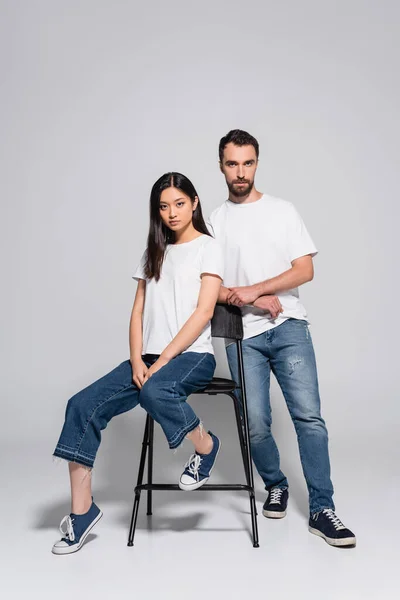 Brunette asian woman looking at camera while sitting on chair near stylish boyfriend on grey — Stock Photo