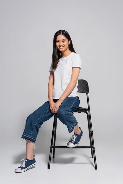 Brunette asian woman in jeans, gumshoes and white t-shirt sitting on chair and looking at camera on grey — Stock Photo