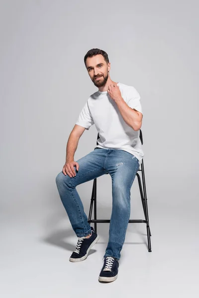 Junger, bärtiger Mann in weißem T-Shirt und Jeans auf Stuhl sitzend, während er in die Kamera auf grau blickt — Stockfoto