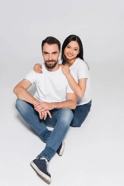 Young interracial couple in white t-shirts and jeans looking at camera while sitting on white — Stock Photo