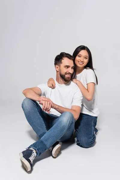 Elegante hombre en blanco camiseta, jeans y gumshoes sentado en blanco cerca de asiático novia - foto de stock