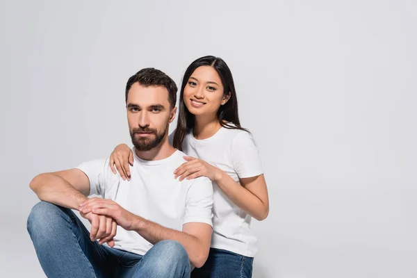 Brunette asian woman touching boyfriend and looking at camera while sitting together isolated on white — Stock Photo