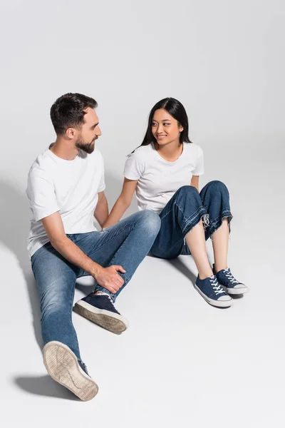 Giovane coppia interrazziale in bianco t-shirt, jeans e scarpe da ginnastica guardando l'un l'altro mentre seduto su bianco — Foto stock