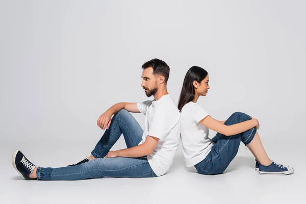 Seitenansicht eines stilvollen gemischtrassigen Paares in weißen T-Shirts, Jeans und Gummischuhen, das Rücken an Rücken auf Weiß sitzt — Stockfoto