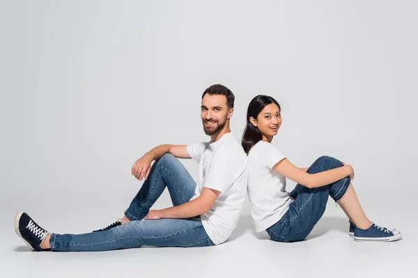 Young asian woman and bearded man in white t-shirts and jeans sitting on white and looking at camera — Stock Photo