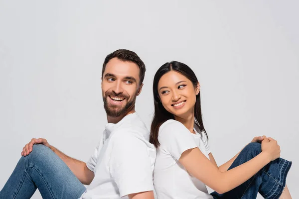 Brunette asian woman and bearded man in white t-shirts sitting back to back isolated on white — Stock Photo