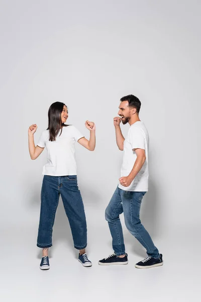 Vue pleine longueur du couple interracial excité en jeans, t-shirts blancs et des chaussures de gomme dansant sur blanc — Photo de stock
