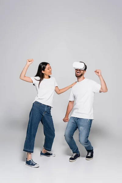 Asian woman in jeans and white t-shirt touching shoulder of excited man dancing and singing in vr headset on grey — Stock Photo
