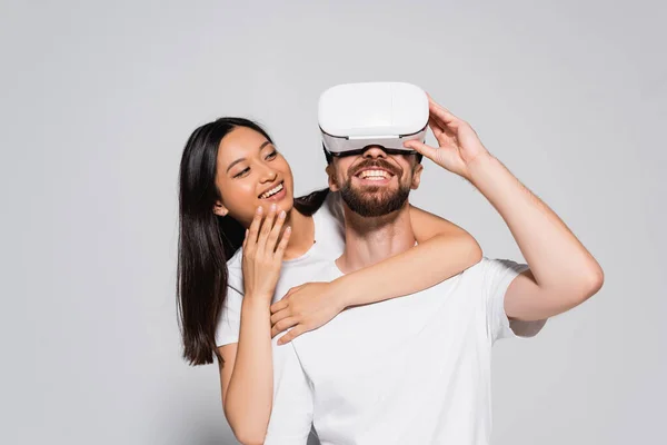 Giovane donna asiatica abbracciare fidanzato eccitato gesturing in vr headset su grigio — Foto stock