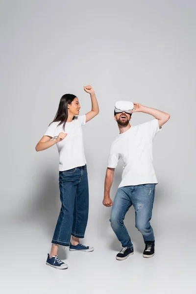 Aufgeregten Mann berührt vt Headset und singt beim Tanzen in der Nähe asiatische Frau auf grau — Stockfoto