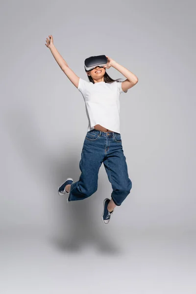 Young asian woman in white t-shirt, jeans and gumshoes jumping with raised hand while using vr headset on grey — Stock Photo