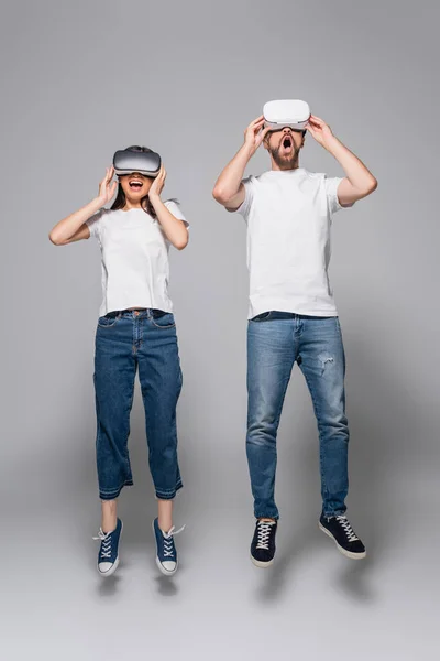 Excited couple in jeans and white t-shirts levitating with open mouths while using vr headsets on grey — Stock Photo