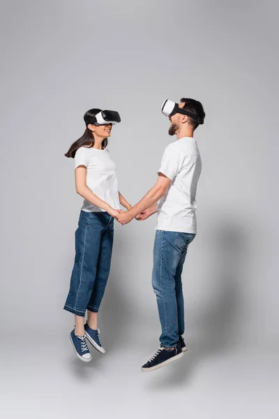 Young couple in jeans and white t-shirts holding hands while levitating in vr headsets on grey — Stock Photo