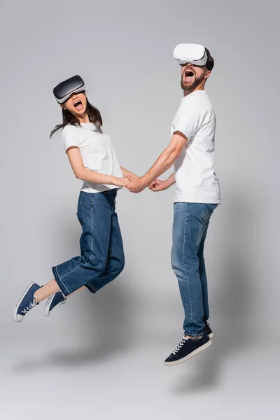 Excited couple in vr headsets, jeans and white t-shirts holding hands while levitating on grey — Stock Photo