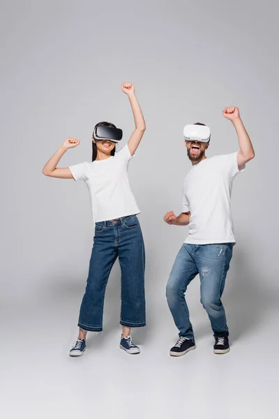 Pareja emocionada en jeans, zapatos de goma y camisetas blancas bailando mientras usa auriculares vr en gris - foto de stock