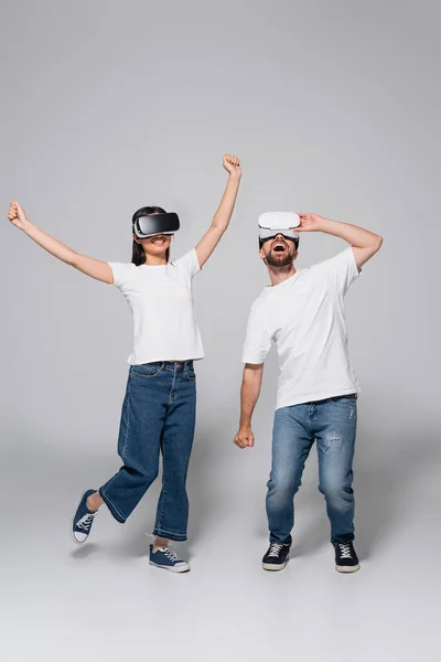 Mujer joven bailando con las manos levantadas cerca del hombre excitado mientras usa auriculares vr juntos en gris - foto de stock