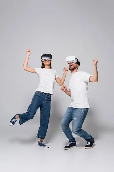 Young, excited couple in vr headsets, jeans and white t-shirts dancing on grey — Stock Photo