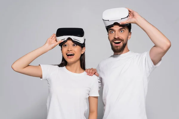 Excited interracial couple in white t-shirts looking away with open mouths while touching vr headsets on grey — Stock Photo
