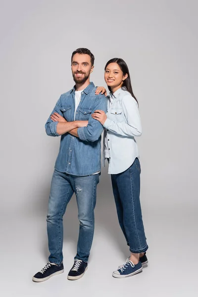 Vista completa de pareja interracial en jeans, camisas de mezclilla y zapatos de goma mirando a la cámara en gris - foto de stock