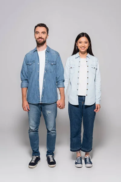 Full length view of interracial couple in denim shirts, jeans and gumshoes holding hands on grey — Stock Photo